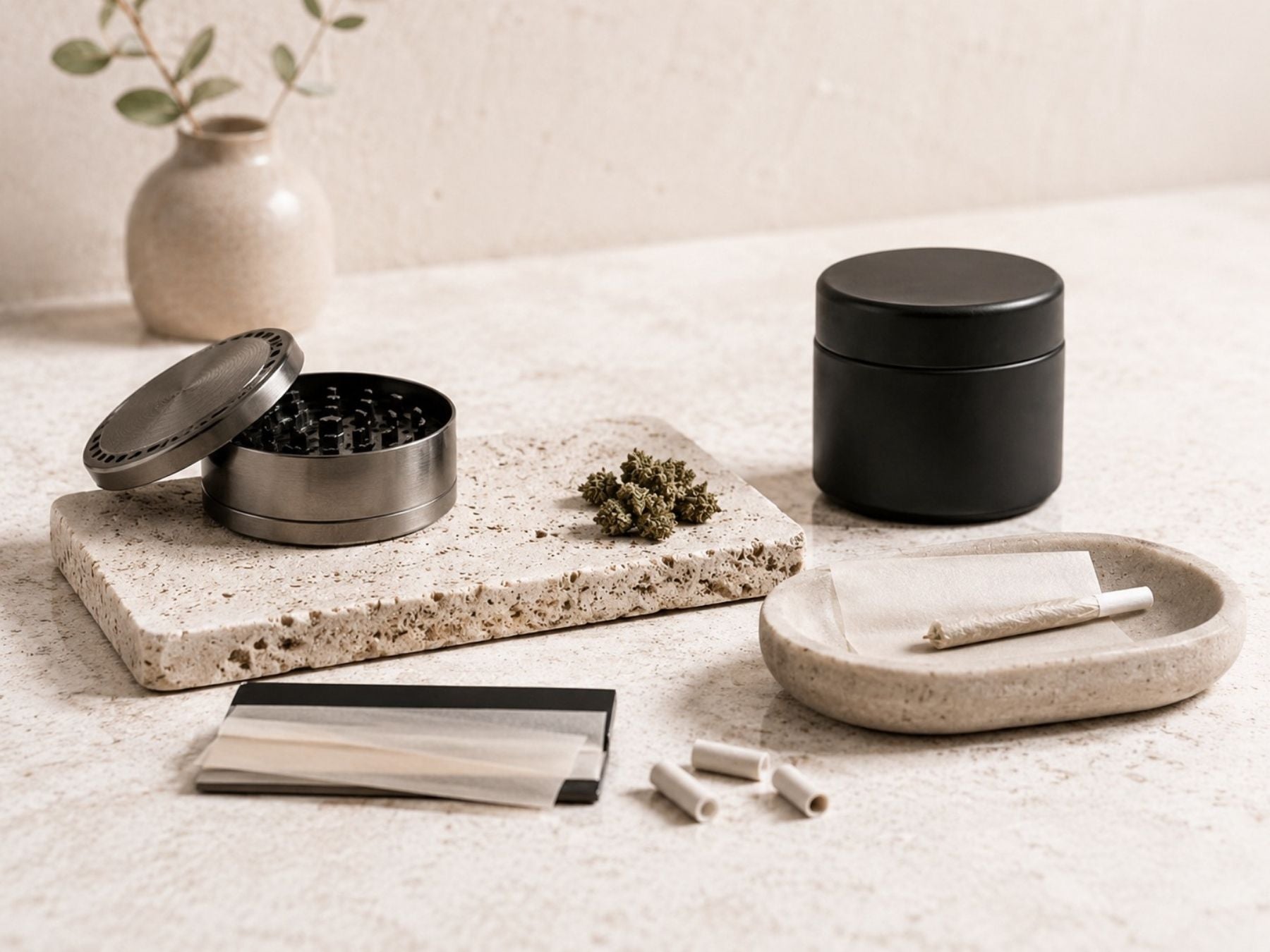 Geöffneter Grinder mit Hanfblüten, Drehpapier, Filtertips und schwarzer Aufbewahrungsdose auf heller Steinplatte
